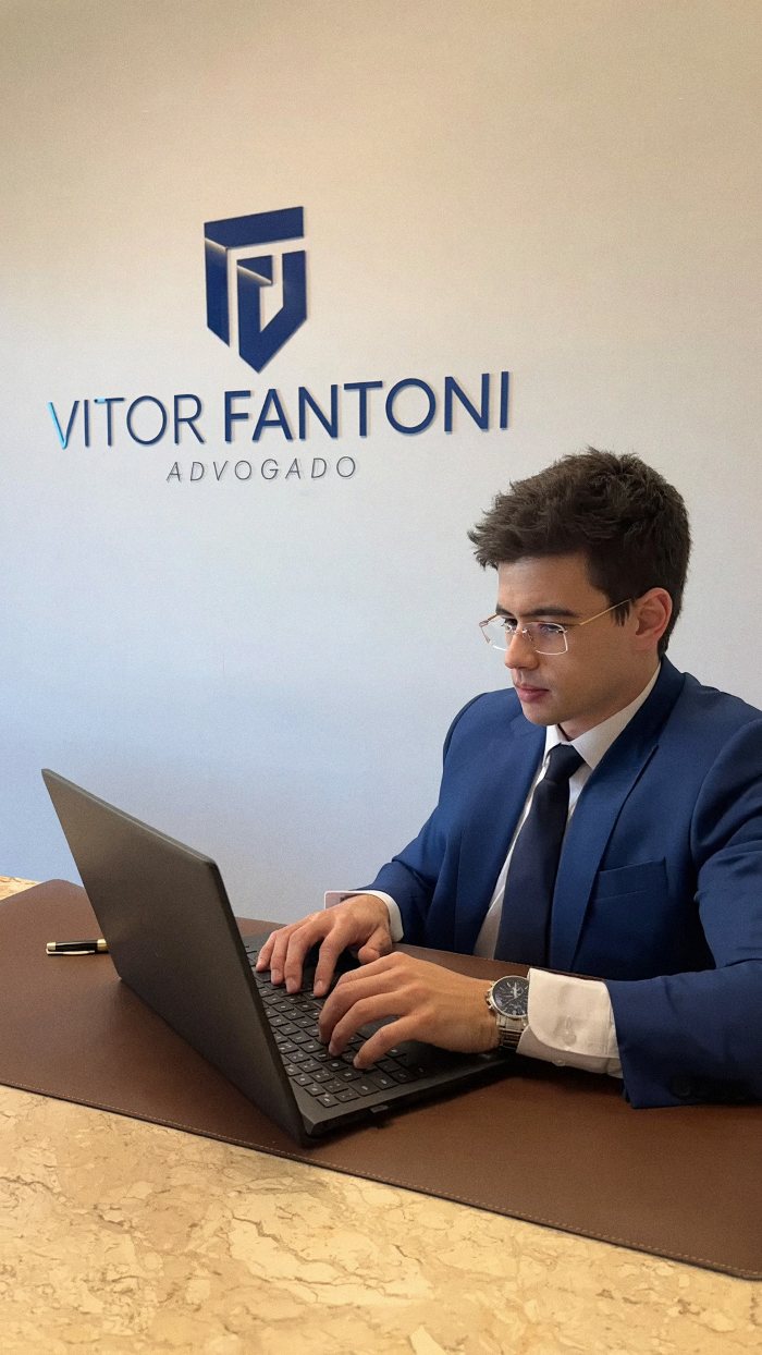 Vitor Fantoni trabalhando no notebook em seu escritório, em frente ao logo Vitor Fantoni Advogado